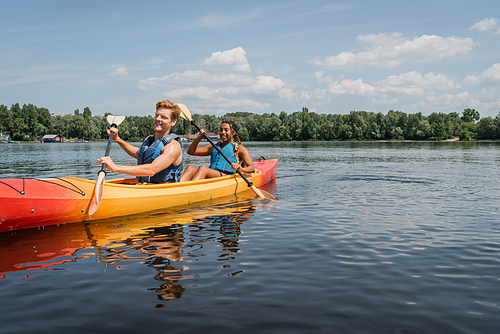 happy and sportive interracial couple in life vests spending vacation on picturesque lake and sailing in kayak with paddles under blue sky with clouds