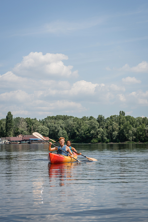 sportive multiethnic couple in life vests paddling in kayak on river with green picturesque bank under blue cloudy sky during recreation weekend in summer