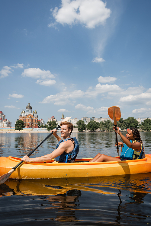 carefree redhead man and pretty african american woman in life vests sailing in sportive kayak along riverside with cityscape and green trees under blue and cloudy sky