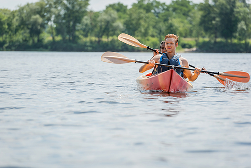 young redhead man in life vest holding paddle and looking away while sailing in sportive kayak with african american woman during summer weekend on river