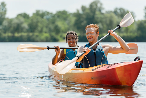 active redhead man and charming african american woman in life vests spending time on river while sailing in sportive kayak on picturesque lake on blurred background in summer