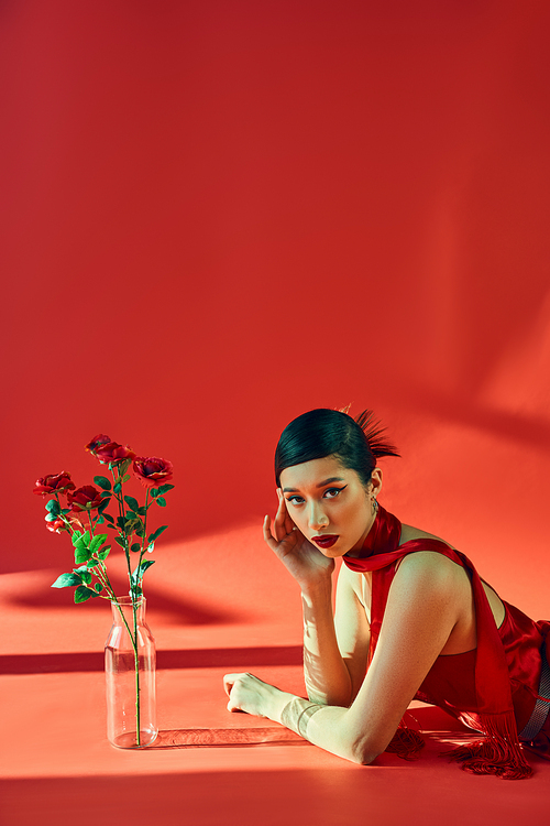 spring fashion concept, young asian woman with brunette hair and bold makeup laying in neckerchief and dress while looking at camera near fresh roses on red background with lighting