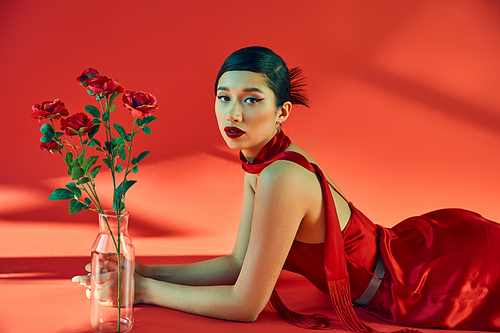 spring fashion concept, young and elegant asian woman with bold makeup, in neckerchief and dress lying near glass vase with roses and looking at camera on red background with lighting