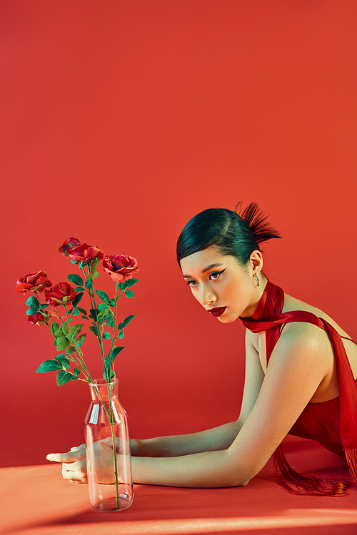 young and elegant asian woman in stylish neckerchief and dress, with bold makeup and brunette hair laying near glass vase with roses on red background with lighting, spring fashion, generation z