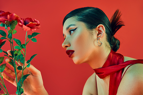 side view of mesmerizing asian woman with brunette hair, bold makeup and trendy hairstyle touching green leaves of roses on red background, generation z, spring fashion concept