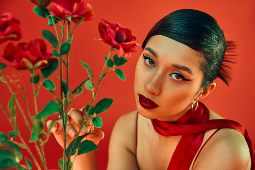 spring style photography, charming asian woman with brunette hair and bold makeup touching green leaves on roses and looking at camera on red background, generation z