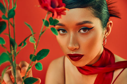 portrait of young asian woman in neckerchief, with bold makeup and expressive gaze looking at camera near flowers on red background, spring fashion photography, generation z