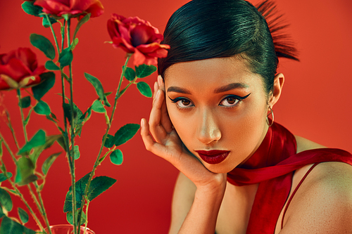 portrait of attractive asian woman with bold makeup, brunette hair and expressive gaze looking at camera near roses on red background, fashionable spring, youthful style