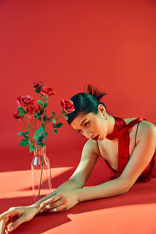 charming asian woman with brunette hair and bold makeup wearing neckerchief and laying on red background with lighting near glass vase with roses, youthful fashion, trendy spring