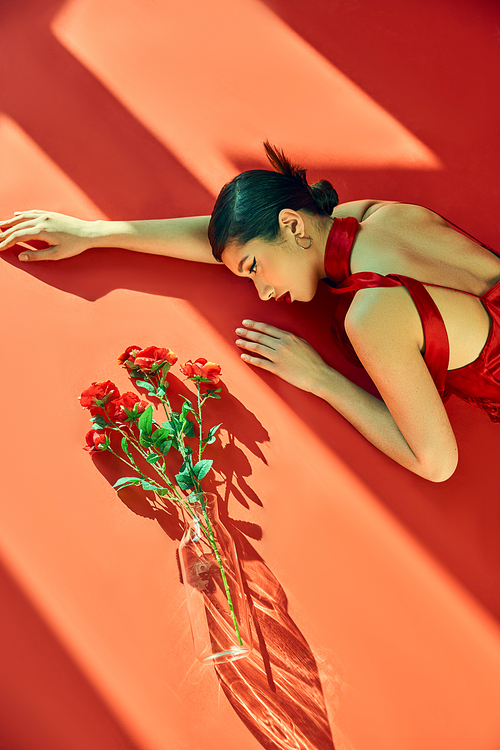 top view of expressive and young asian woman with brunette hair, in neckerchief and dress lying in lighting near glass vase with roses on red background, generation z, spring fashion photography