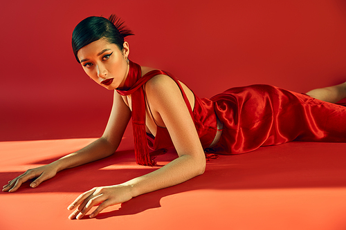 seductive asian woman with trendy hairstyle and bold makeup, in elegant dress and neckerchief lying and looking at camera on red background with lighting, youth culture, stylish spring concept