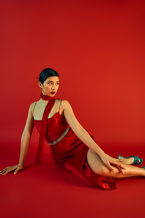 charming asian woman with brunette hair and bold makeup looking away while sitting on red background in neckerchief and strap dress, generation z, spring fashion concept