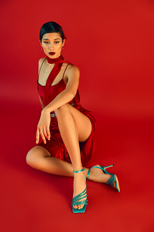 full length of elegant asian woman in stylish spring outfit sitting in stylish pose on red background, brunette hair, strap dress, neckerchief, turquoise sandals, generation z fashion