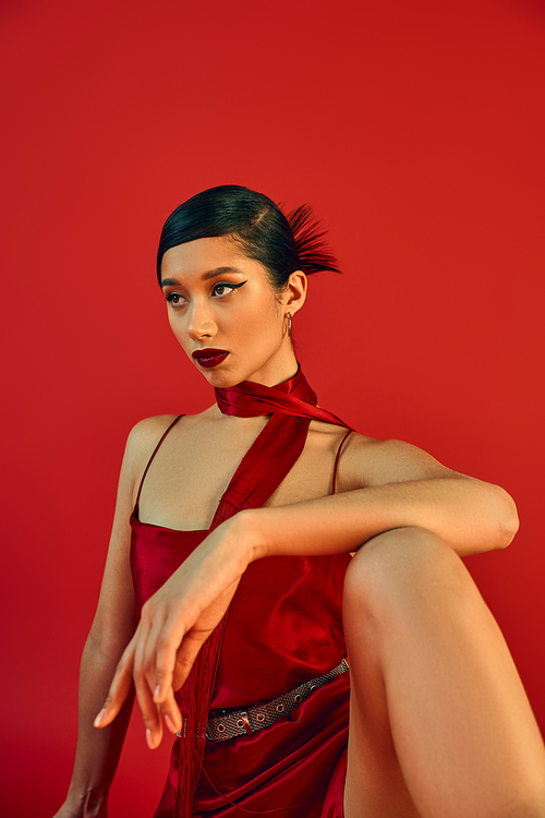 youthful and confident asian woman with bold makeup, brunette hair and expressive gaze looking away and posing in neckerchief and elegant strap dress on red background, spring fashion photography