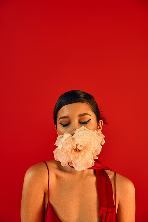 spring fashion photography, portrait of appealing asian woman with brunette hair, in stylish neckerchief, posing with closed eyes and fresh peony in mouth on vibrant red background