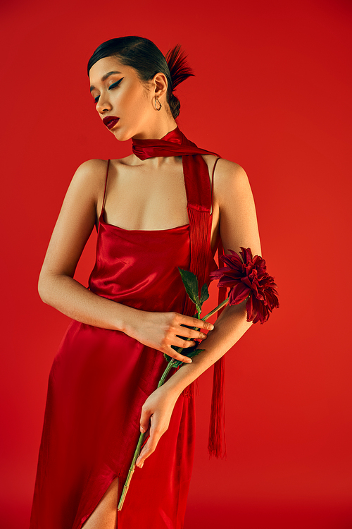 youthful and sensual asian woman with bold makeup and brunette hair, in glamour dress and neckerchief posing with burgundy peony on red background, trendy spring, gen z fashion