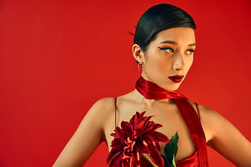 charming asian woman with bold makeup, brunette hair and expressive gaze, in strap dress and neckerchief holding burgundy peopny and looking away on red background, spring fashion photography