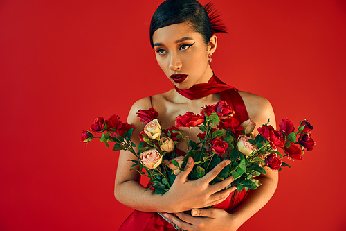 elegant asian fashion model with bold makeup, brunette hair and expressive gaze holding bouquet of roses and looking away on red background, generation z, trendy spring