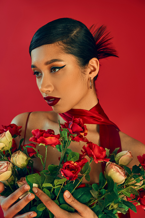 spring style concept, youthful fashion, graceful asian woman with brunette hair, bold makeup and trendy hairstyle posing in neckerchief while holding fresh roses on red background