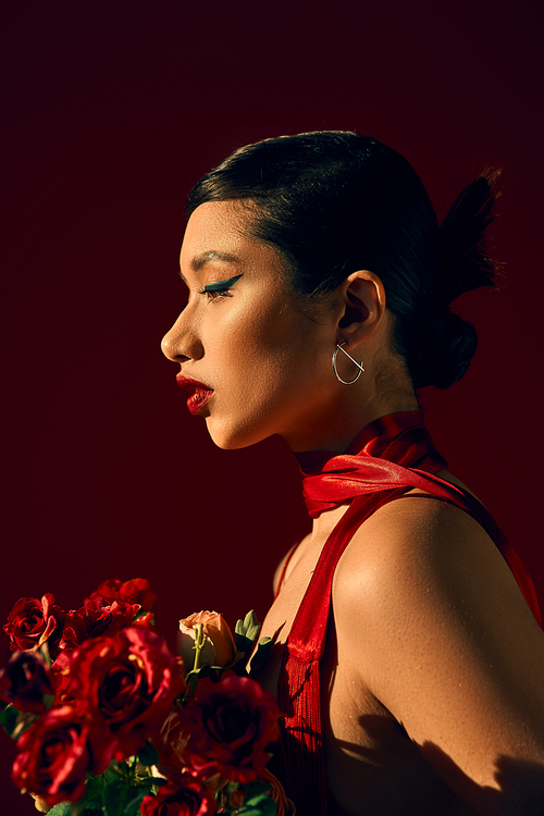 profile of charming asial woman with brunette hair and bold makeup, in silver earring and red neckerchief holding bouquet of roses on dark background, spring fashion photography