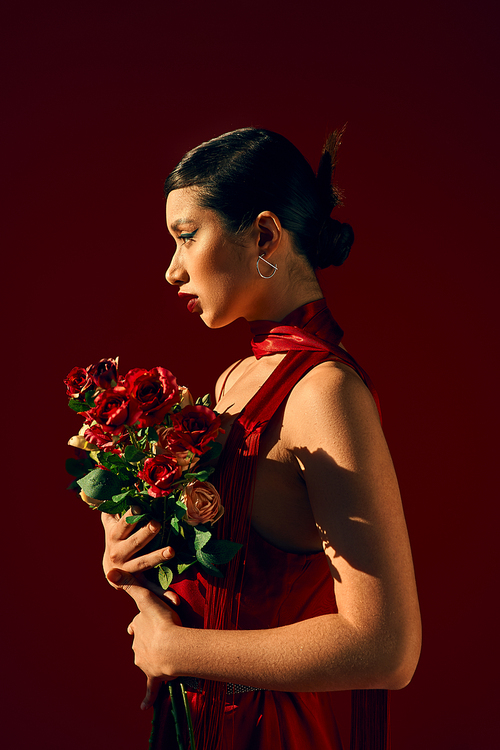 side view of mesmerizing asian fashion model in red neckerchief, with brunette hair and bold makeup posing with bouquet of roses on dark background, trendy spring, fashion shoot