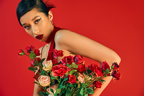 youthful and mesmerizing asian woman with bold makeup, brunette hair and seductive gaze looking at camera near bouquet of fresh roses on red background, trendy spring, generation z