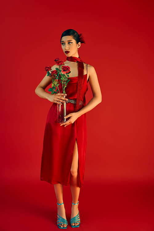 full length of youthful and elegant asian woman in stylish strap dress and neckerchief, with bold makeup and brunette hair standing with roses on red background, trendy spring, generation z