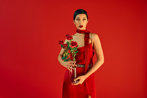 gen z fashion, stylish spring concept, charming asian woman in elegant dress and neckerchief holding glass vase with roses and looking away on red background