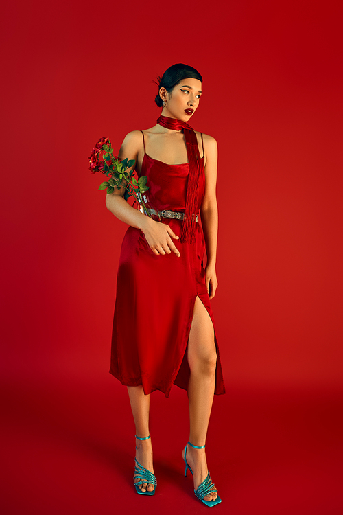 full length of appealing asian woman in spring outfit standing with flowers on red background, elegant strap dress, neckerchief, turquoise sandals, fashion shoot