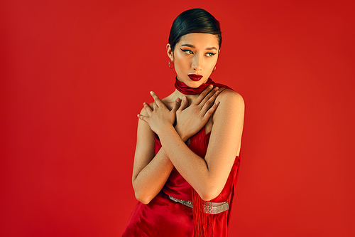 young asian woman with brunette hair and bold makeup holding hands on chest and looking away while posing in neckerchief and dress on red background, fashionable spring