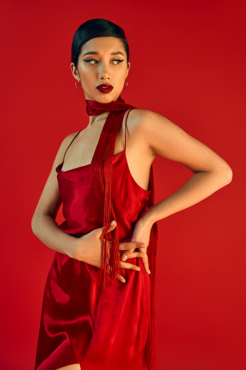 young and fashionable asian woman with bold makeup and brunette hair wearing elegant strap dress and neckerchief while posing with hands on hip on red background, spring fashion concept