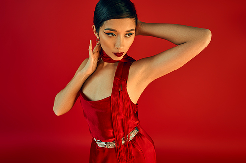 youthful and expressive asian woman with brunette hair and bold makeup, wearing neckerchief and elegant dress, looking away on red background, gen z fashion, stylish spring concept