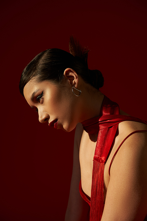 profile of youthful asian woman with brunette hair, bold makeup and silver earring posing in neckerchief and strap dress on dark red background with lighting, spring fashion concept