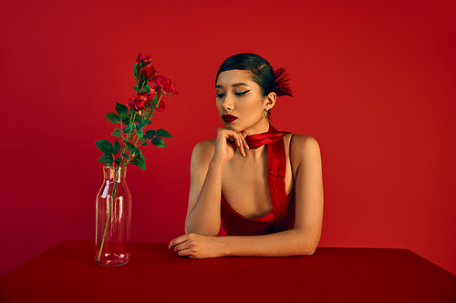 romantic asian woman in dress and neckerchief, with brunette hair and bold makeup sitting at table neat glass vase with roses on red background, fashion photography, stylish spring concept