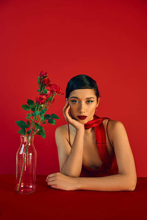 trendy spring, youthful fashion, pretty asian woman with bold makeup and brunette hair sitting in neckerchief near glass vase with roses and looking at camera on red background