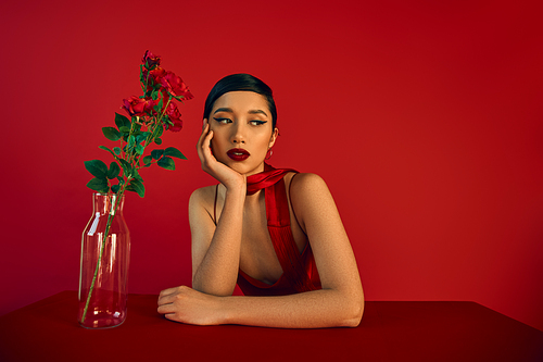 alluring asian woman with brunette hair and bold makeup, in elegant dress and neckerchief sitting at table near glass vase with fresh roses on red background, spring fashion concept