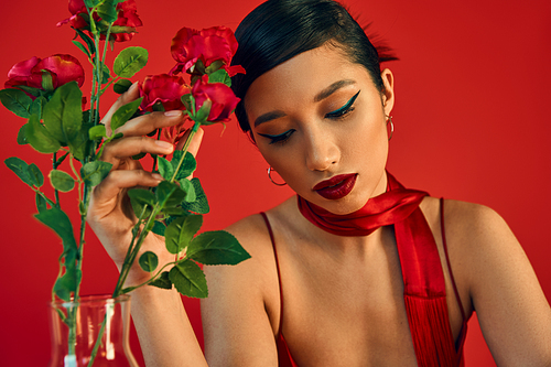 sensual and dreaming asian woman with bold makeup, brunette hair and stylish neckerchief touching fresh roses on red background, spring style, fashion photography