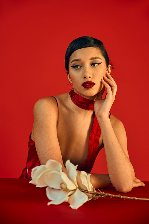 dreamy asian woman with brunette hair and bold makeup holding hand near face and looking away near white orchid on red background, spring fashion, strap dress, stylish neckerchief