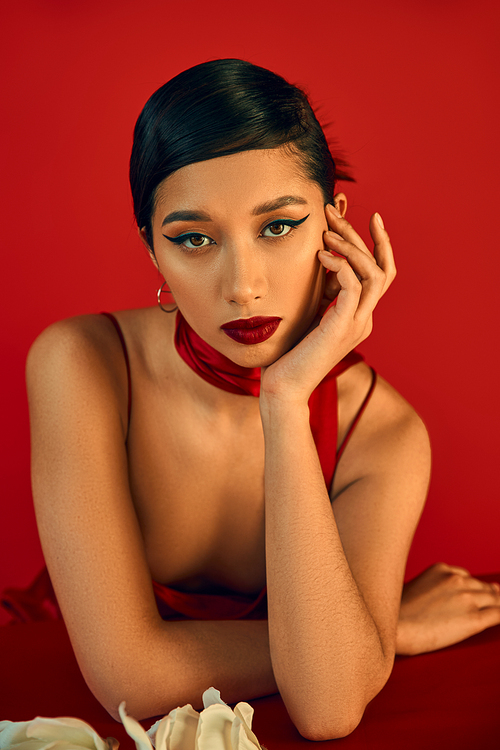 portrait of charming and trendy asian woman in strap dress and neckerchief, with brunette hair and bold makeup sitting with hand near face and looking at camera on red background, spring fashion