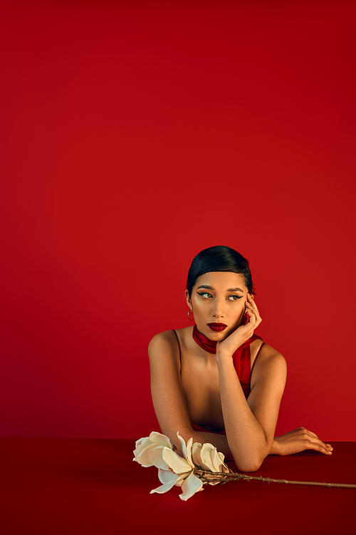 pensive asian woman in stylish neckerchief, with brunette hair and bold makeup sitting at table near while orchid, holding hand near face and looking away on red background, trendy spring concept