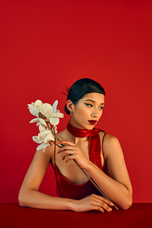 young and beautiful asian woman with brunette hair and bold makeup holding white orchid while sitting at table in neckerchief and strap dress on red background, trendy spring, gen z fashion