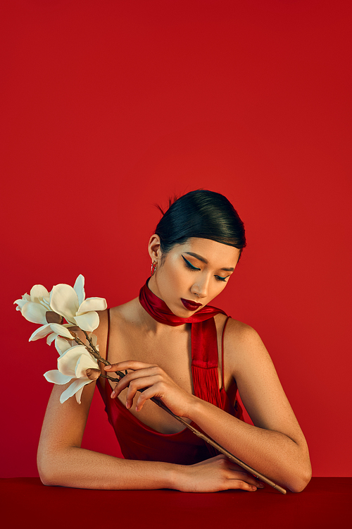 youthful fashion, trendy spring concept, charming asian woman in neckerchief and strap dress sitting at table with white blooming orchid on red background