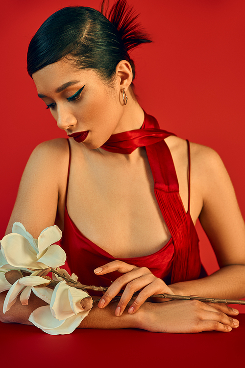 elegant and young asian woman in trendy dress and neckerchief, with brunette hair and bold makeup sitting at table and looking at white orchid on red background, gen z fashion, trendy spring concept