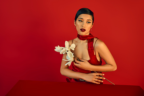 sensual and brunette asian woman with bold makeup, in elegant dress and neckerchief sitting at table with white blooming orchid on red background, generation z, trendy spring concept