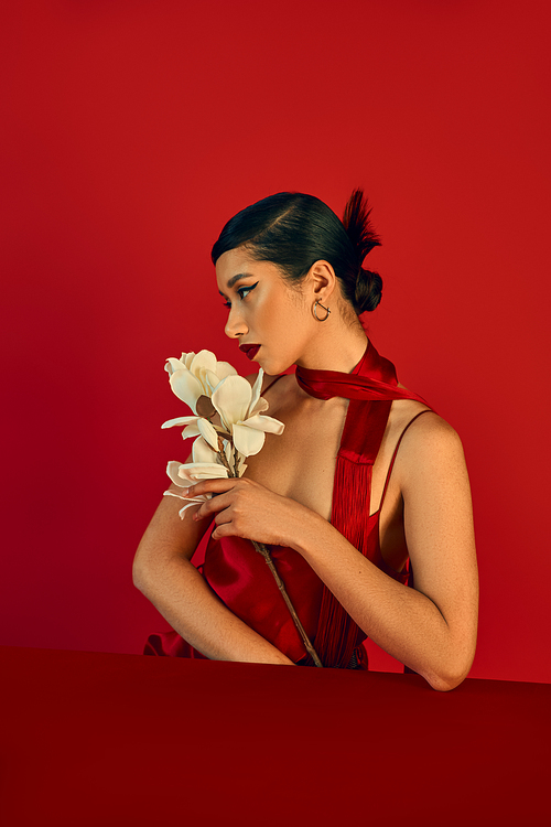 spring fashion concept, young asian woman sitting at table with white orchid and looking away on red background, brunette hair, bold makeup, strap dress, neckerchief, gen z style