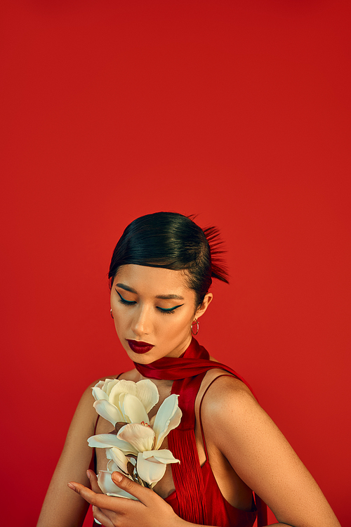 youthful and charming asian fashion model in strap dress and neckerchief, with brunette hair and bold makeup looking at white blooming orchid on red background, trendy spring, fashion shoot