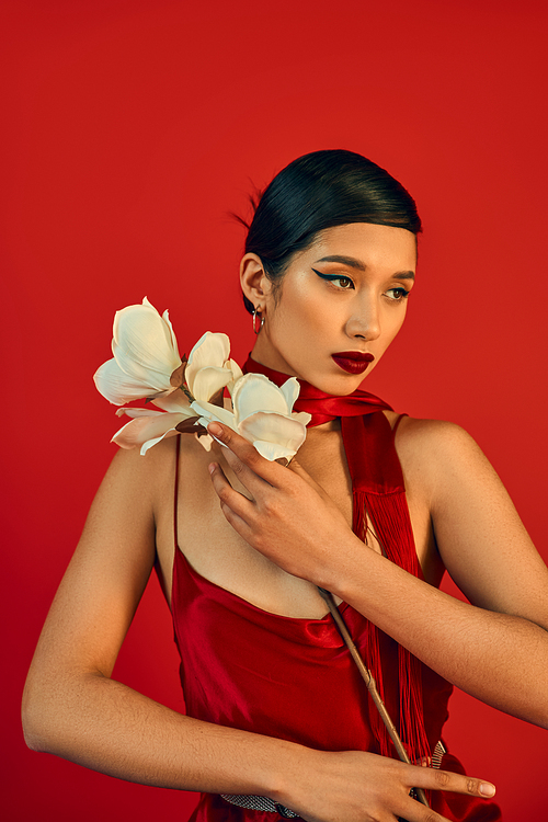 romantic asian woman in spring outfit holding white orchid and looking away on red background, brunette hair, bold makeup, strap dress, neckerchief, generation z