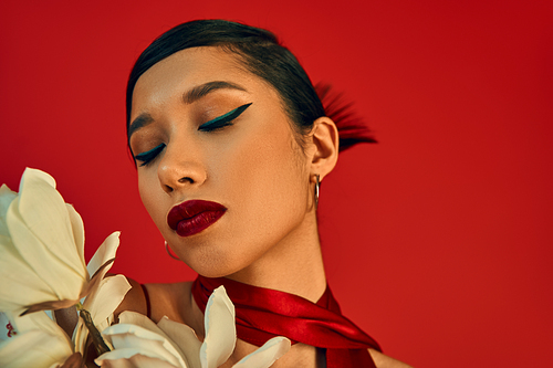 portrait of sensual asian fashion model with brunette hair, bold makeup and trendy hairstyle posing with closed eyes near white blooming orchid on red background, spring fashion, generation z