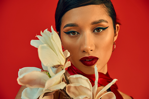 portrait of youthful and mesmerizing asian woman with brunette hair, bold makeup and expressive gaze looking at camera near while blooming orchid on red background, spring fashion photography