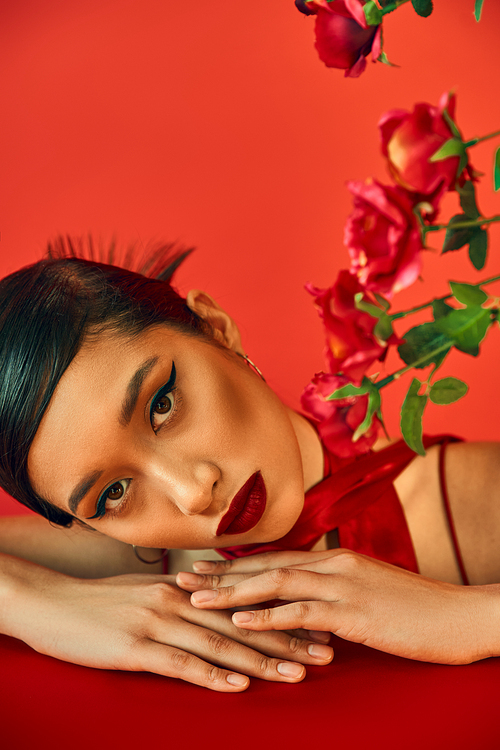 portrait of young and appealing asian fashion model with bold makeup and brunette hair looking at camera while laying on table near roses on red background, stylish spring, fashion shoot